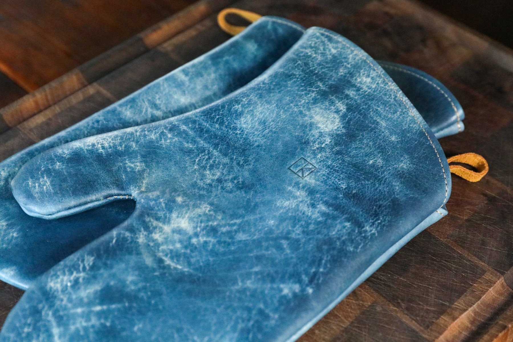 A pair of blue leather oven mitts with a distressed, marbled texture and small yellow handles, resting on a dark wooden cutting board.
