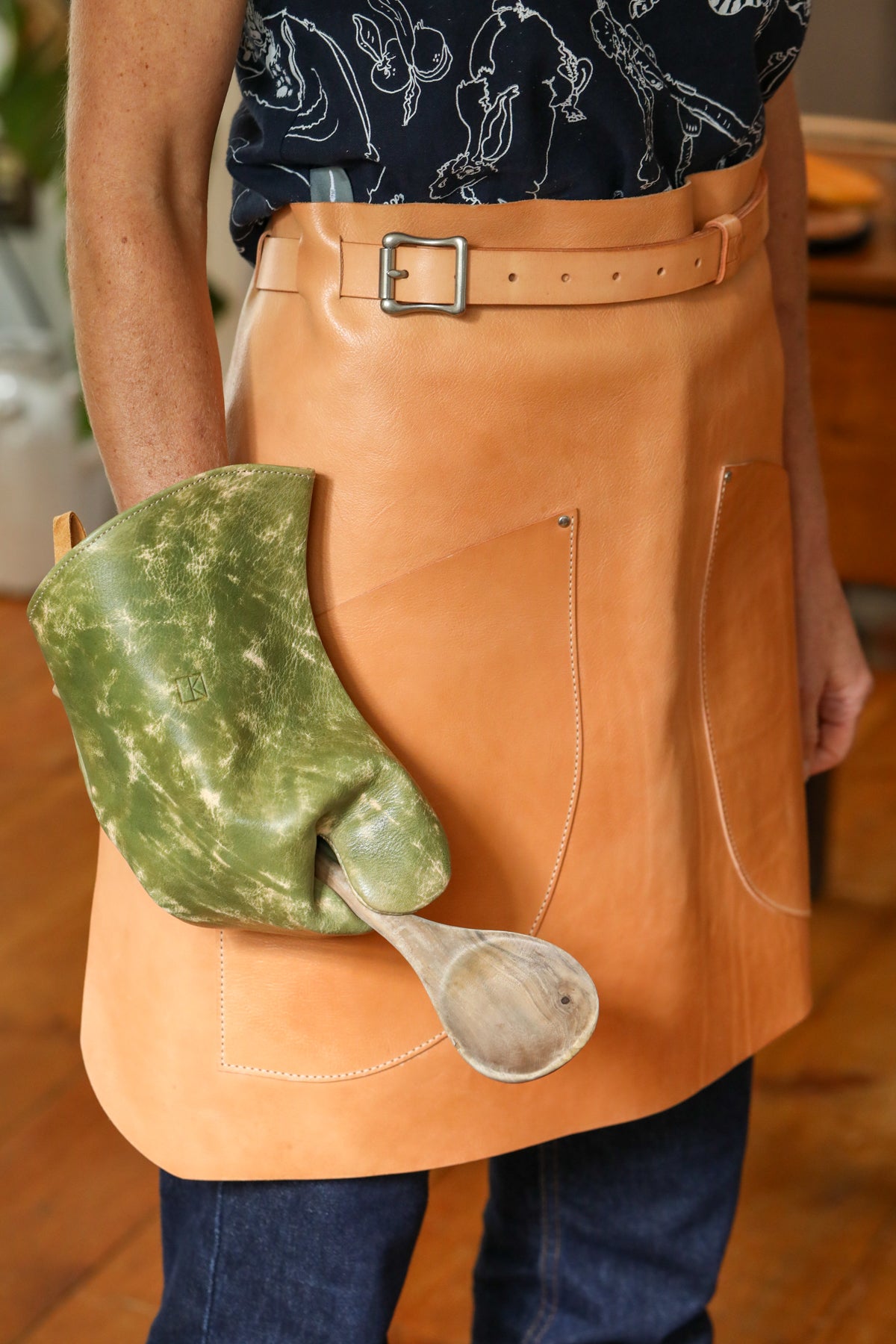 A person wearing a tan leather apron with a belt and front pockets, holding a green marbled cloth or bag in one hand and a wooden spoon in the other, against a warm indoor background.