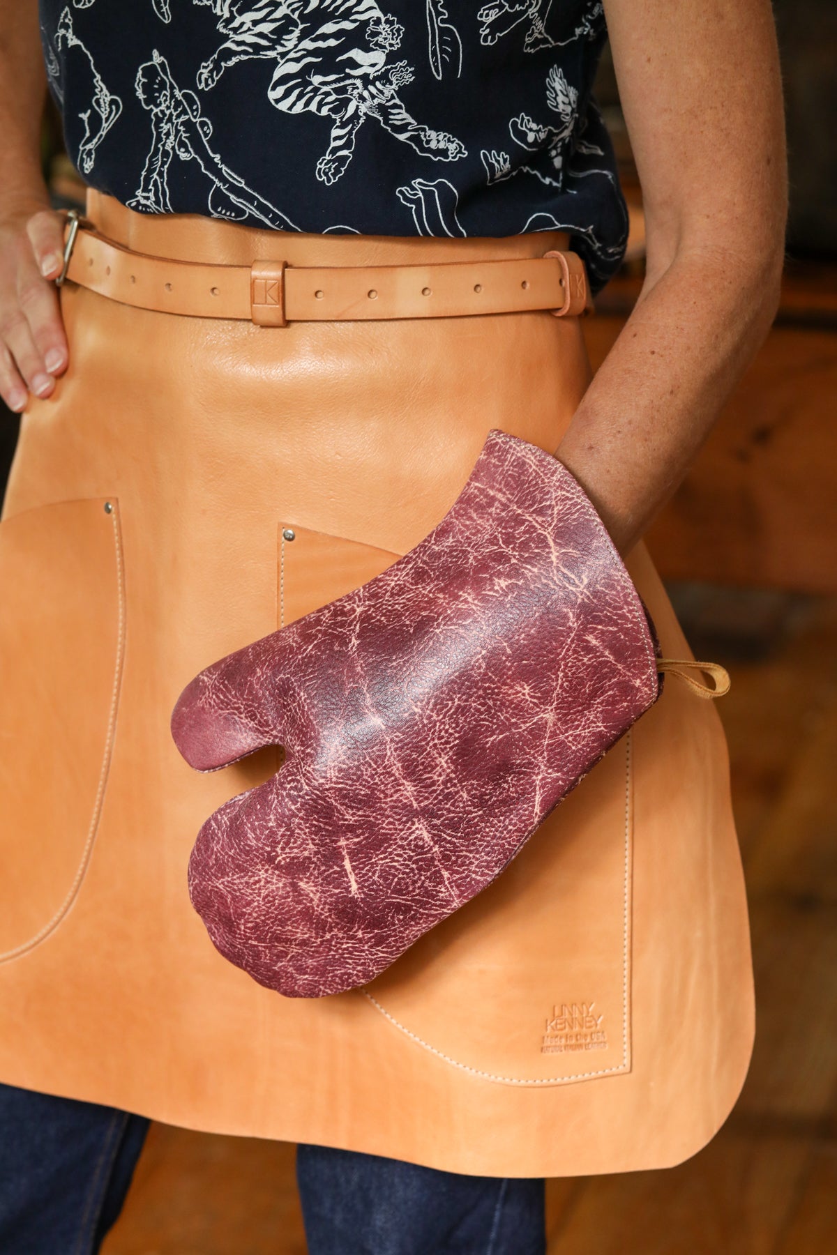 A person wearing a tan leather apron and holding a distressed burgundy leather oven mitt. The apron has a belt and front pockets, and the person is wearing a dark patterned top and jeans.